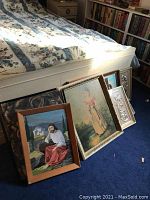 Multiple framed paintings leaning against a bed with visible subjects: Jesus in a white shirt and red robe, a woman in period dress, and an ornate metallic frame design
