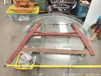 Antique buck saw frame shown on a round glass table with measuring tape indicating length. Frame is painted red with a rusty saw blade attached at bottom.