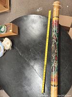 Full length image of bamboo rainstick on black table next to measuring tape showing length approx 38 inches. Painted tribal designs visible.