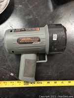 Photo showing a grey handheld cordless rechargeable spotlight with a black lens cap and a pistol grip handle, placed on a black surface with a measuring tape below.