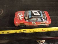 Front-side view of the unopened Dale Earnhardt Jr. NASCAR racecar chocolate tin, showing length against tape measure.