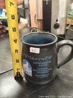 Black ceramic mug measuring about 4.5 inches tall with Department of Justice text in multiple languages including Greek, English, French, and Inuktitut. Features Nunavut polar bear logo. Photo shows tape measure for scale.