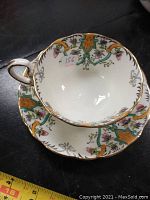 Close-up of bone china tea cup with floral and geometric pattern, gold rim and handle, showing condition and design details with measuring tape beside.