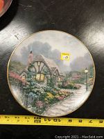 Front view of the Thomas Kinkade collector plate depicting a cottage with gardens and glowing windows, accompanied by a measuring tape showing size.