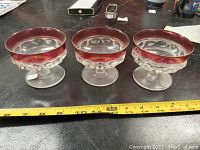 Three vintage glass dessert cups with red colored rims and clear bases with embossed designs, placed side by side on a surface with measuring tape showing approximate size.