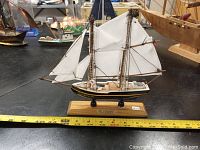Front side view of the vintage handmade wooden ship model "The Bluenose" with sails and wooden base with name plaque, next to measuring tape showing approx 12 inches length.