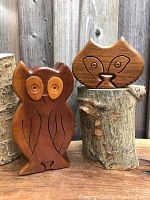 Photo showing owl and cat wooden puzzle boxes side by side on wooden stumps, highlighting size and basic shape.