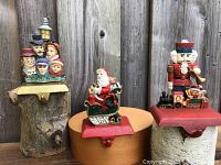 Three resin Christmas stocking hangers on cast iron bases including Santa in sleigh, carolers, and nutcracker figures.