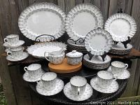 Full set arranged on wooden surface against fence showing plates, cups, saucers, trays, creamer and sugar bowl