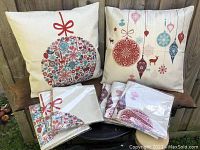 Two assembled pillow cases displayed on a wooden surface, one with a large ornament design and one with hanging ornaments, plus sealed packaged pillow cases of the same designs underneath.