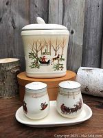 Full view of the Hallmark cookie jar featuring Santa in sleigh with reindeer, two salt and pepper shakers with winter illustrations on a matching tray, arranged on a wooden surface against a wood panel background.