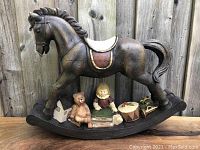 Front view of large brown rocking horse figurine with accompanying small toy figurines including a teddy bear, a doll, books, a house, a drum, and a horn, placed on wooden surface against a wooden fence background.
