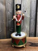 Front view of the wooden nutcracker drummer music box standing on a wooden surface against a wooden plank background. Shows full figure with details on hat, face, uniform, and drum base.