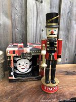 Nutcracker standing on a wooden surface beside its holiday-themed box with a character face mug visible inside the box.