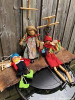 Three vintage hand crafted wood marionette puppets displayed on a wooden bench against a wooden fence: Pinocchio, Anne of Green Gables, and a pirate. Wooden control bars and visible strings are shown.