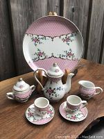 Miniature porcelain tea set arranged on wooden surface showing tray in background with teapot, sugar bowl with lid, creamer, and two teacups with saucers in front.