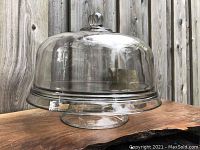 Glass cake tray with pedestal base and clear dome lid, photographed outdoors on wooden surface against wood fence.