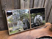 Two Christmas-themed light-up wall hangings displayed outdoors showing winter village scenes in wooden frames.