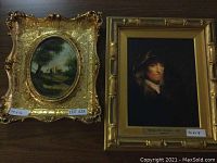 Two framed art pieces side by side on wooden surface, showing both the landscape in ornate oval gold frame and portrait in rectangular bamboo style gold frame