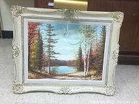Framed landscape painting showing a lakeside scene with an attached overhead light and cords hanging down.
