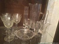 Full view of the lot in display cabinet showing pitcher, vase, basket dish, beaded rim dish and eight glasses with etched Cornflower pattern