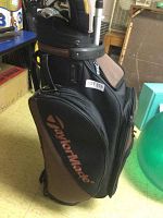 Side view of TaylorMade golf bag with visible clubs inside, located indoors on a floor with various other items