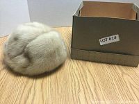 Side angle view of the light beige fur hat showing its plush texture and fullness. Box labeled 'LOT 614' nearby.