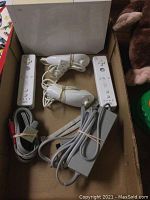 Top view of two white Nintendo Wii Remote controllers, two Nunchuk controllers, power adapter and AV cables bundled with rubber bands inside an open box.
