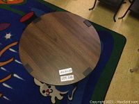 Top view of round wooden tabletop showing wood grain and four black metal corner brackets with sticker showing dimensions and lot number.