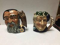 Front view of Royal Doulton Toby jugs Merlin and Bacchus, showing detailed facial expressions and glossy ceramic finish.