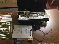 Shows silver trumpet in open black soft case with various accessories including mouthpieces and valve oil bottles on the table around it.