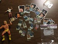 Photo showing two Hulk Hogan wrestling action figures, multiple small sealed wrestling trading cards, loose small wrestling cards, photos, and a pin/button on a wooden table.