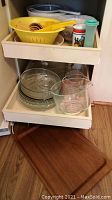 View of two pullout shelves with yellow plastic nested colanders and scoops on top shelf, glass measuring cup, pie plates, and baking dishes on lower shelf, teak serving tray on floor.