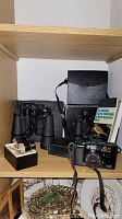 Shelf containing Canon AF 35mm camera, two pairs of binoculars in cases, a guidebook, and Elite opera glasses.