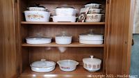 Shelf with white divided French white bakeware with lids and various round and rectangular casseroles with lids in floral and fruit patterns.