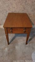 Front view of the Beaucraft Elmira wood end table showing drawer with ornate metal handles and tapered legs.