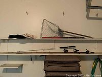 Shelf holding three fishing poles with reels, fishing nets, fishing umbrella, and three hats.