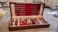 Wooden canteen box open displaying multiple rows of silver plated knives, forks, spoons, and flatware with floral handles and red felt lining.
