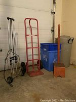 Rubbermaid garbage pail, two blue recycling bins, wooden step stool with tall handle, red metal hand cart, and silver metal folding hand cart arranged near garage door.