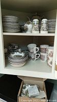 Photo showing upper and lower shelves inside cupboard with stacks of dinner plates, salad plates, bowls, mugs, salt and pepper shakers.