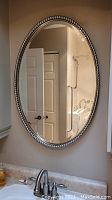 Full front view of the oval wall mirror hung above a bathroom sink, showing the mirror reflecting part of the bathroom interior.