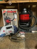 Photo showing Shop-Vac unit with hose and attachments in front of its filled original box alongside Bissell SpotLifter handheld shampooer with power cord coiled.