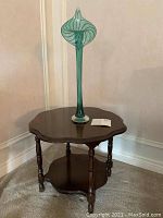 End table with dark stained wood, scalloped edges, turned legs, and lower shelf shown with vase on top.