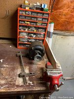 Photo of the workbench showing a red painted metal vise attached to the edge, black General Electric AC motor behind it, and a red metal sorting box with plastic drawers filled with hardware items on the wall behind.