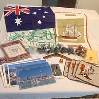 Overview of the lot showing the Australian flag, linen tea towels with printed designs, three stuffed koalas, two stuffed echidnas, two stuffed kangaroos, embroidery pattern packet, laminated placemats with Aboriginal designs, and New Zealand postcards.