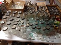 Full lot of vintage clear and green glass canning jars with metal lids, rings and boxes of lids, displayed on table.