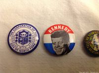 Close-up photo of three pins including 'The Corporation of the City of North York', 'Kennedy' political button, and 'Vacation' themed button