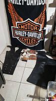 Overview photo showing two pairs of black leather chaps on the floor in front of a large Harley-Davidson flag and several collectible items.