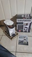Photo of Baby Brezza Formula Pro Advanced dispenser unit, box with printed product image, and instruction manual on tile floor.