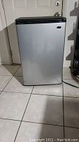 Front view of a silver mini refrigerator with black top handle in a tiled room under artificial light.